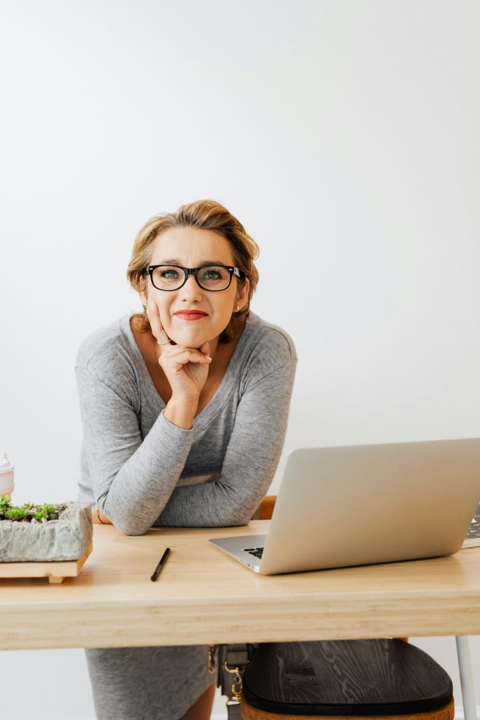 pexels-photo-5240021-5240021 Smiling woman sitting at desk with laptop and plants, ideal for modern office settings.