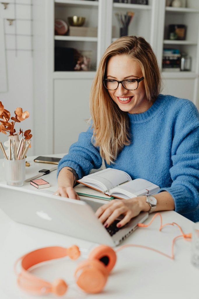 pexels-photo-5908728-5908728 A cheerful woman in a blue sweater working remotely with a laptop in a cozy home setting.