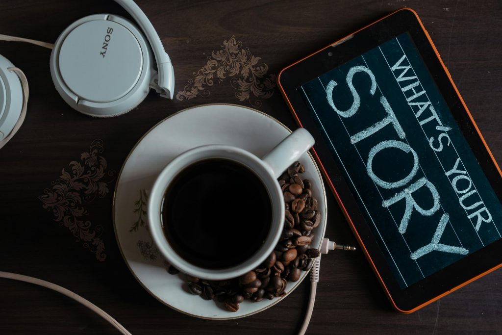 pexels-photo-1749303-1749303 Flat lay of coffee cup, headphones, and tablet with 'What's Your Story' on screen.